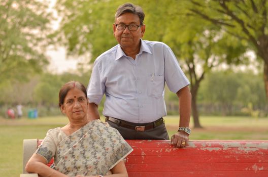 Happy looking retired senior Indian man and woman wearing saree couple smiling sitting on a red park bench in an outdoor setting in New Delhi, India