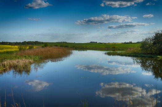 Tranquil pond with reflections of surrounding fields and cloudy blue sky in a picturesque rural landscape