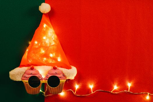 Greeting Season concept.Santa Claus hat with star light and glasses that decoration with Christmas cupcake on red and green background