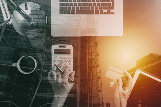 Internet shopping concept.Top view of hands working with calculator and laptop and credit card and tablet computer on dark wooden table background with buildings exposure