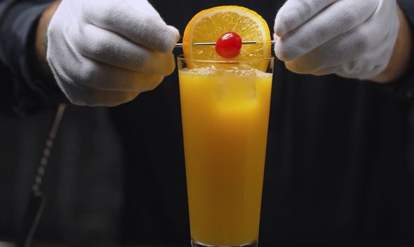 Preparing a Tequila Sunrise Cocktail. Close up bartenders hands garnish it with orange and cherry on skewer.