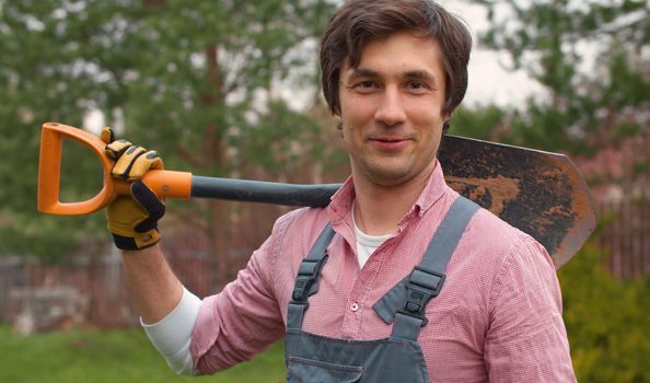 Portrait of a gardener with a shovel on his shoulder. Smiling young caucasian man in overalls in the garden at spring day