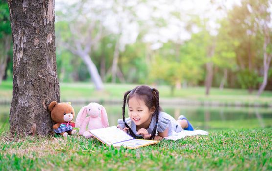 cute little girl  reading a book while lying with a doll in the park