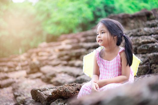 Little asian girl with yellow umbrella sitting on th rock of historic site in Thailand