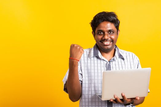 Closeup Asian happy portrait young black man excited holding laptop computer clenching fists and raising a hand for winner sign celebrating his success, studio shot isolated on yellow background