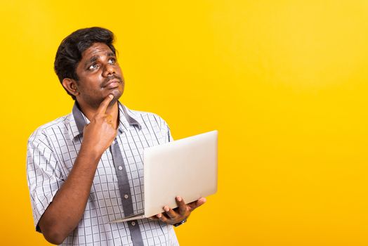 Closeup Asian happy portrait young black man holds a laptop computer hand finger handle on the chin and thinking question looking away, studio isolated on yellow background