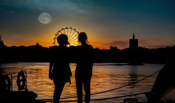 silhouettes of two young girls with ferris wheel in the back at sunset in Malaga port, Spain