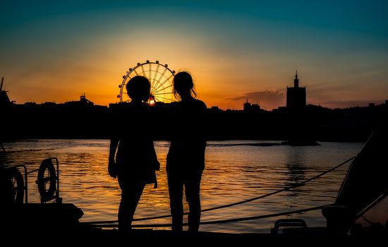 silhouettes of two young girls with ferris wheel in the back at sunset in Malaga port, Spain