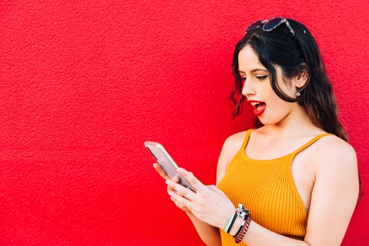 Woman looking at her phone with mouth wide open in red background.