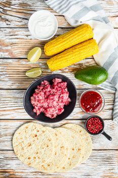 Cooking Mexican taco ingredients with minced organic beef meat in black bowl , corn, calsa over white textured wooden table, top view