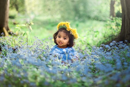 A small girl with dandelion wreath is sitting on field of forget me not blue flowers