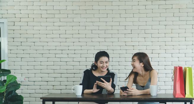 Two Asian women who are laughing and attractive are friends, talking about coffee in a coffee shop. Beautiful and confident women use a tablet to shop online together.