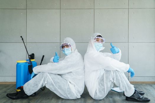 Two young men wearing protective clothing and medical masks are tired from work and sitting on the floor. Cleaning services are very popular after the spread of coronavirus or COVID-19.