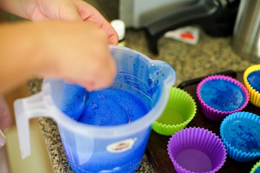 Melting soap in a plastic mug and adding colors and fragrances before pouring into colorful silicon molds to make home made melt and pour soap or blue colored cupcake muffins during the coronavirus lockdown