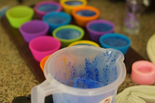 Melting soap in a plastic mug and adding colors and fragrances before pouring into colorful silicon molds to make home made melt and pour soap or blue colored cupcake muffins during the coronavirus lockdown