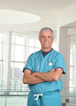 Smiling male doctor in scrubs with a stethoscope around his neck and his arms crossed. Vertical format 3/4 torso view in modern looking medical facility.