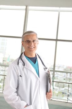 Middle aged doctor with stethoscope and clip board standing in front of large window in modern medical facility