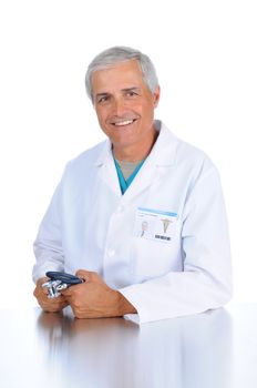 Smiling middle aged doctor seated and holding his stethoscope in both hands. Man is wearing a lab coat and scrubs in vertical format over a white background.