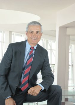 Middle aged businessman standing in modern office lobby leaning o none knee. Vertical format with man in front of large window and smiling at the camera.