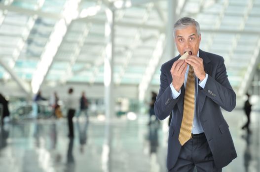 Middle aged Businessman on the go eating a sandwich in Airport Concourse, horizontal format with unrecognizable people in the background.