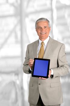 A mature businessman holding a tablet computer with blue screen facing forward. Easy to replace screen to add your own graphic. Vertical format against blurred modern office interior.