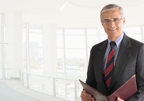 A mature businessman with a leather folder in a modern office building. The background is out of focus and high key. Horizontal format with copy space.