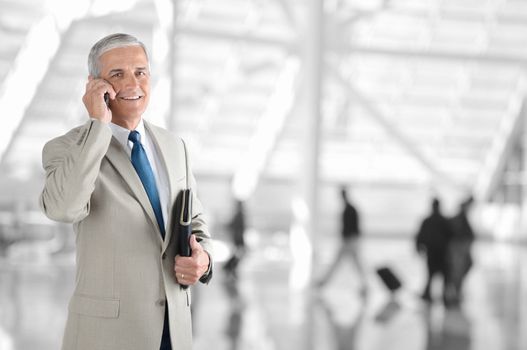 A mature businessman on his cell phone in an airport terminal. The background is blurred with unrecognizable people. 