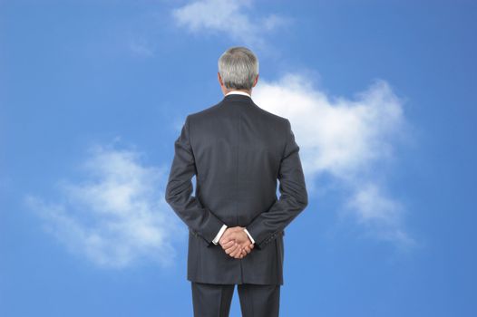 Middle aged Business with hands behind back standing in front of blue cloudy sky
