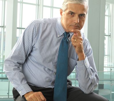 Middle aged businessman with serious expression seated with hand on chin in modern office.