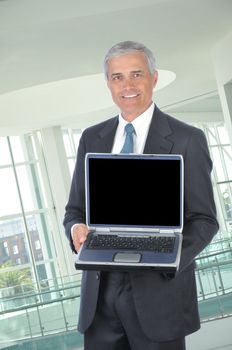 Smiling Middle aged Businessman in dark suit standing with laptop computer open and facing the camera in office setting