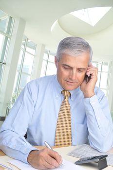 Middle aged Businessman seated at his desk talking on phone and taking notes in office setting