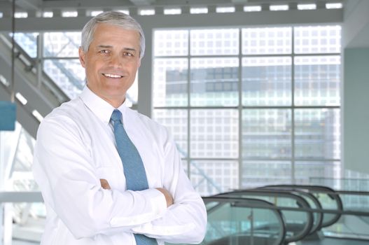 Smiling Middle aged Businessman with arms crossed Standing in Building Lobby with esclaator in background