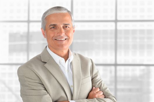 Portrait of a middle aged businessman in front of office window. Man is smiling and has his arms folded. Horizontal format.