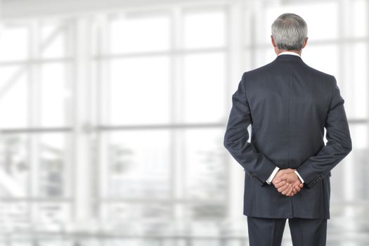 Portrait of smiling senior businessman standing looking out of office window with his hand behind his back.