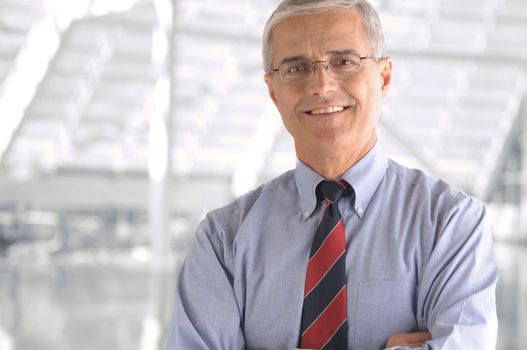 Business man portrait in modern office building. Middle aged man is smiling at the camera and has his arms folded. Closeup head and shoulders only.