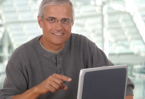 Middle aged man sitting and pointing at laptop computer in modern office setting. Man is smiling and casually dressed looking at camera. Horizontal format.