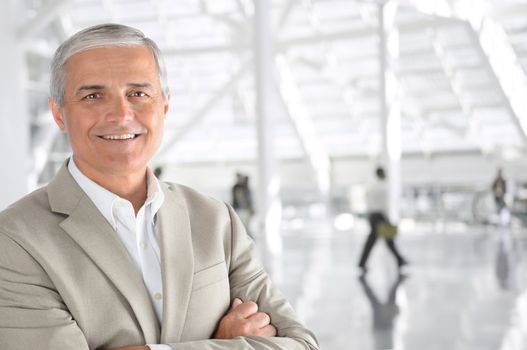 Closeup of a mature businessman with his arms folded in an airport concourse. The man is smiling at the camera with blurred passengers in the background.
