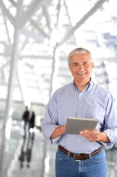 A mature businessman in the lobby of a modern office building. The casually dressed man is smiling and there are blurred people in the background.