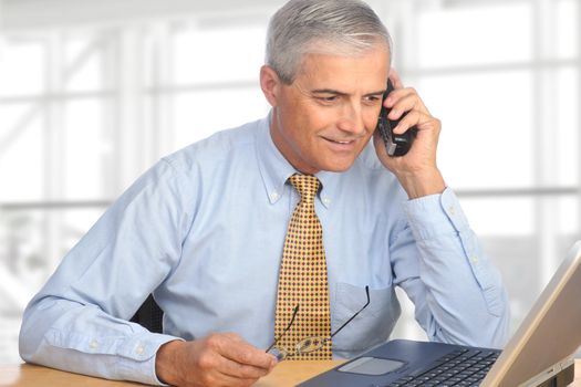 A mature salesman talking on the telephone. The man is seated in a modern office with a laptop on his desk in front of a large bank of windows.