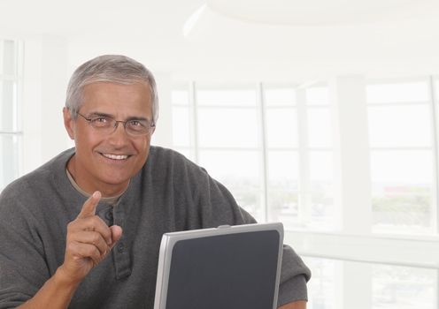 A smiling casually dressed mature businessman seated behind a laptop computer pointing towards viewer.. The man is in front high key office window.