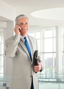 Portrait of a middle aged businessman standing in a modern office talking on a cell phone.. Man is holding a small binder and smiling at the camera.