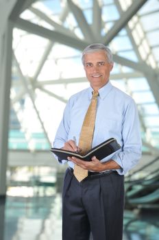 Smiling Middle aged Businessman Standing in Building Lobby and Writing in Notebook 