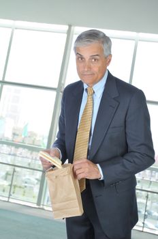 Middle aged Businessman in office building holding brown bag and sandwich with quizzical look on his face.