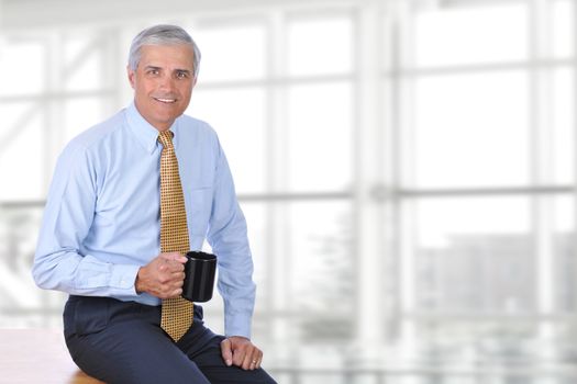 A mature businessman sitting on the corner of his desk in a modern office with a coffee mug in his hand. Horizontal format with copy space. 
