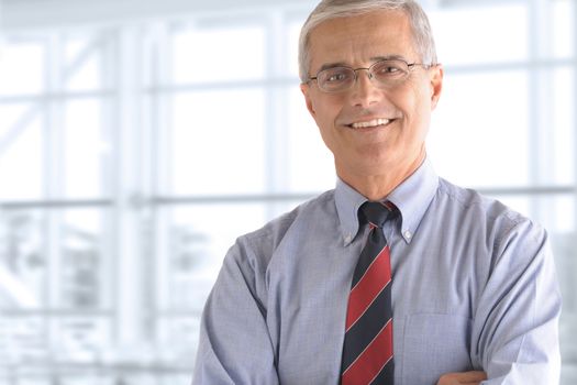 Portrait of a mature businessman standing in front of a large office window. The man is smiling and has his arms crossed.