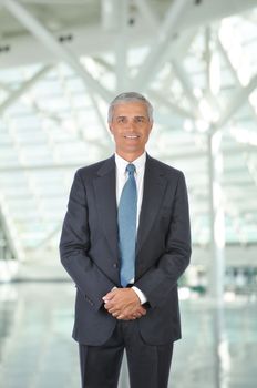 Middle aged businessman standing in modern office lobby with his hands in front of body. Vertical format with man smiling at the camera.