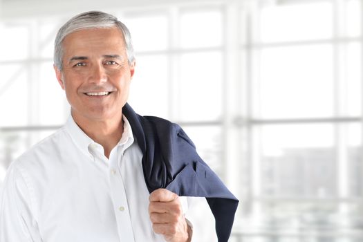 Portrait of smiling senior businessman standing against office window background while looking at camera, with his coat over his shoulder