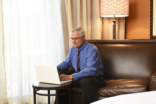 A senior businessman seated at the desk in his hotel room and working on his laptop computer. Horizontal format.