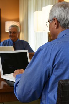A senior businessman seated at the desk in his hotel room and working on his laptop computer. Vertical format with the mans reflection in the mirror.