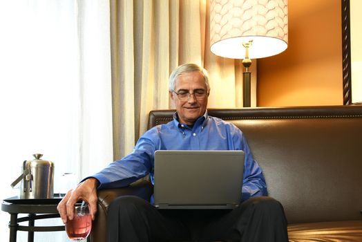 Mature Businessman sitting on the couch in his hotel room holding a drink while working on his laptop. Business travel concept.